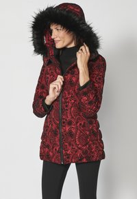 Crvena puffer jakna s uzorkom, s kapuljačom od umjetnog krzna, zatvaranjem na patentni zatvarač i prilagođenim manžetama. Sadrži složene crne paisley dizajne na tkanini.