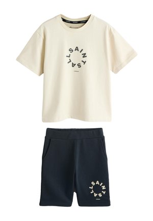 Nastolatek stoi i się uśmiecha, ubrany w beżowy t-shirt i czarne szorty z okrągłymi wzorami tekstu, białe skarpetki i białe sneakersy na jednolitym tle.