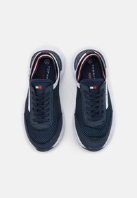 Sneaker in rete blu navale con accenti bianchi, parte superiore testurizzata e lacci piatti. Presenta un design semplice e un'etichetta con logo sul lato.