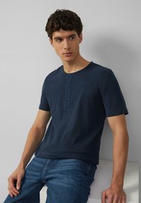 Marineblauwe kortarm henley t-shirt van gestructureerde stof met een drieknops sluiting en ronde hals, gecombineerd met een blauwe spijkerbroek.