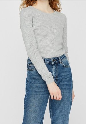 Vrouw die een lichtgrijze, lange mouwen, gebreide trui draagt en hoge taille, blauwe denim jeans, staand met één arm licht gebogen.