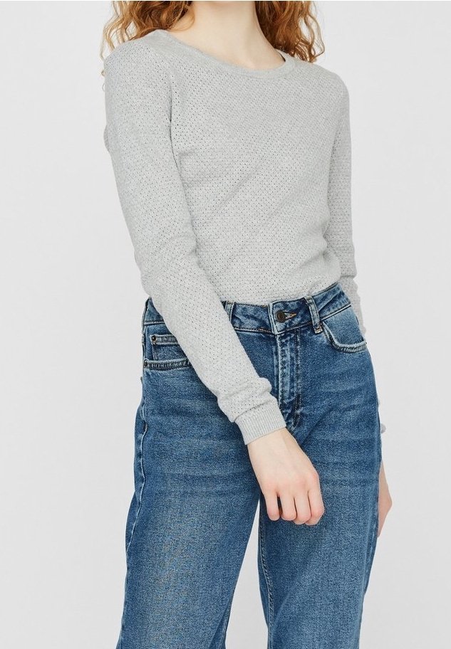 Femme portant un pull en maille gris clair à manches longues et un jean bleu en denim taille haute, debout avec un bras légèrement plié.