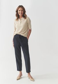 Beige short-sleeve polo shirt with a collar and three buttons, paired with dark navy, relaxed-fit pants; model wearing beige heeled mules.