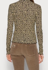 Zwarte blouse met lange mouwen en gele bloemenpatroon. Zacht materiaal, aansluitend ontwerp. Paar bruine broeken met logo-patch op de achterzak.