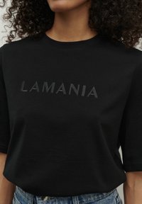 Černé tričko s krátkým rukávem, které má vyvýšený nápis "LAMANIA" v tón v tónu. Vyrobeno z měkké bavlny s volným střihem.
