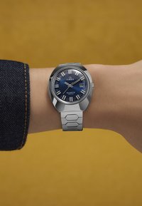 Ezüst Timex quartz óra kék számlappal, pálcikajelzőkkel és dátumfunkcióval, egy hatszögletű lánccal a csuklón. Háttér sárga.