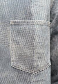 Svetlo modrá denimová tkanina s jemnou textúrou, obsahujúca náprsné vrecko s vystuženým šitím, šikmým otvorom a vybledlým vzhľadom.