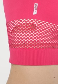 Rosa Sportoberteil mit Mesh-Belüftungseinsatz und "B DRY"-Label darüber, das auf der Bauchregion einer Person gezeigt wird.