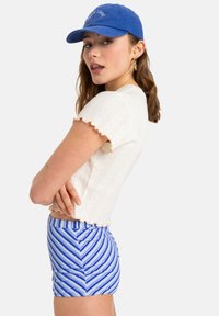 White textured crop top with ruffled sleeves, paired with blue and white striped high-waisted shorts. Blue cap with logo.