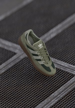 Sneakers en suede vert et mesh avec trois bandes blanches et semelle en caoutchouc marron. Comprend un col et une languette rembourrés, logo sur la languette et sur le côté.