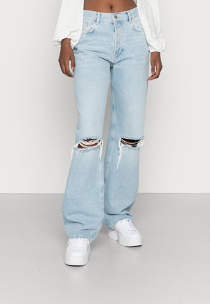Persoon draagt lichtblauwe gescheurde spijkerbroek met grote kniebeschadigingen, witte platformsneakers en een witte lange mouwen top met pofmouwen.