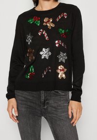 Schwarzer Pullover mit festlichen Paillettenmotiven von Zuckerstangen, Stechpalmen, Schneeflocken und Lebkuchenfiguren auf der Vorderseite. Lange Ärmel.