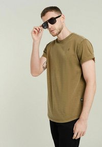 T-shirt in cotone verde oliva con maniche corte, scollatura rotonda e un piccolo logo. Il modello indossa occhiali da sole neri, posando su uno sfondo neutro.