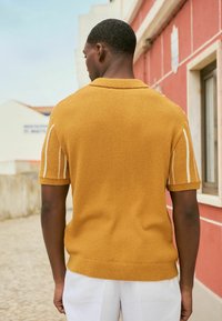 Polo de punto de manga corta en color mostaza, con tejido texturizado, cuello acanalado y rayas blancas en los hombros. El modelo está de pie frente a un fondo colorido.