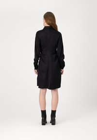 Črna satenska obleka slim fit kroja z dolgimi rokavi, ovratnikom in detajliranjem v pasu. Gladka tekstura z diskretnim vzorcem in dolžino do kolen.