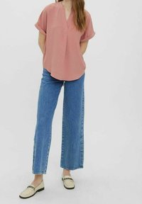 Blouse rose clair à manches courtes avec col en V, associée à un jean en denim bleu à jambes larges et des chaussures beige à enfiler avec des accents noirs.