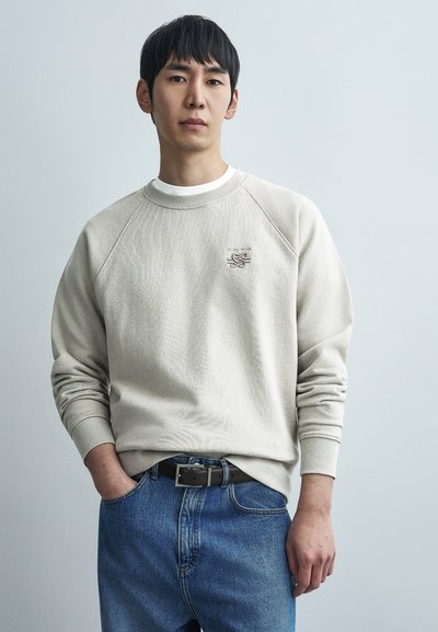 Μπεζ φούτερ με λαιμόκοψη crewneck, με μανίκια raglan, υφασμάτινο με υφή και μικρό κεντημένο λογότυπο. Συνδυασμένο με μπλε τζιν και μαύρη ζώνη.