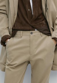 Beiga byxor med en mjuk textur och knäppning, tillsammans med en brun mockaskjorta med knappar, bärs under en ljusbeige kappa.