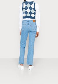 Jeans en denim bleu clair avec une coupe droite, poches arrière et une étiquette en cuir au niveau de la taille. Portés avec un gilet en laine de couleur bleu marine à motifs argyle.