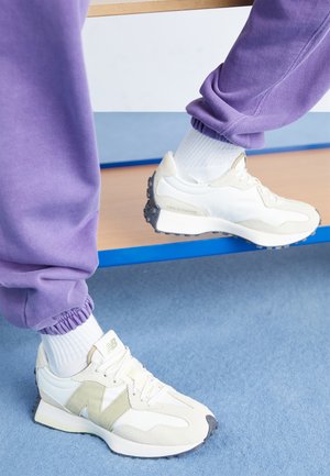 Białe i beżowe sneakersy New Balance noszone z białymi skarpetkami i fioletowymi dresami na niebieskim dywanie obok drewnianej ławki.