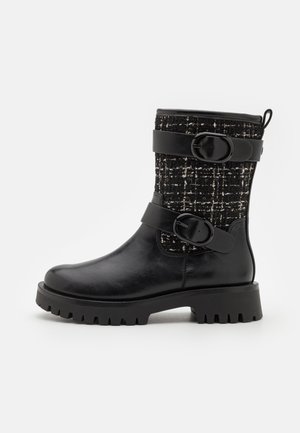Botte d'ankle en cuir noir avec une tige en tissu texturé noir et blanc, dotée de deux larges sangles et de boucles rondes. Semelle en caoutchouc épaisse.