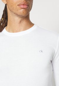 Biela dlhá rukávová tričko vyrobené z mäkkého materiálu, s okrúhlym výstrihom a malým šedým logom "ck" na ľavej hrudi.