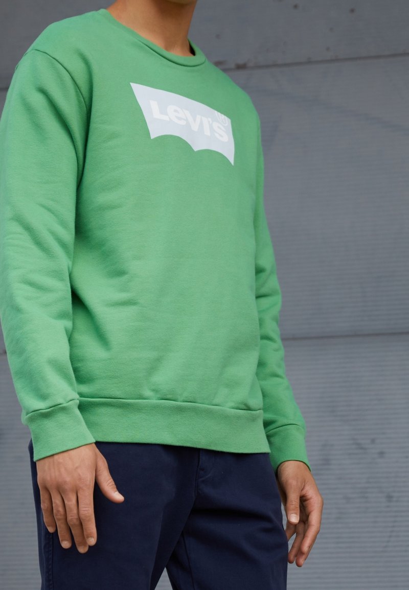 Personne portant un sweatshirt Levi's vert fluo et un pantalon bleu foncé, se tenant devant un arrière-plan en métal ondulé gris.