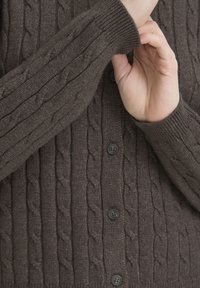 Osoba dopasowująca mankiet brązowego, rozpinanego swetra z grubym splotem i czarnymi guzikami.