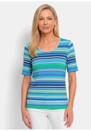Femme aux cheveux blonds et raides portant un t-shirt à manches courtes et col rond avec des rayures horizontales bleues et vertes, associé à un pantalon blanc.