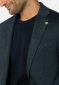 Donkerblauwe blazer van textuurstof, met een enkele knoopsluiting en een met een klep. Gecombineerd met een marineblauwe crew neck shirt.