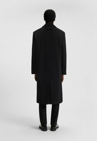 Cappotto in lana nera con colletto strutturato, apertura singola e vestibilità su misura, abbinato a pantaloni neri e scarpe lucide.
