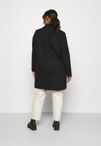 Donna con i capelli scuri raccolti in una coda di cavallo, che indossa un lungo cappotto nero, pantaloni crema con orli sfrangiati e stivali neri, in piedi con la schiena rivolta verso un muro bianco.