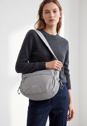 Femme portant un pull gris foncé et un pantalon bleu marine, portant un sac bandoulière gris clair avec le logo "GMV" sur la poche avant.