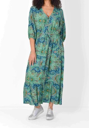 Vrouw draagt een lange blauwgroene paisley jurk met driekwart mouwen en witte sneakers.