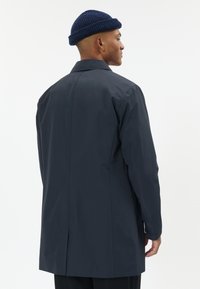 Vyras dėvi tamsiai mėlyną paltą ir tamsiai mėlyną megztą kepurę, žiūrintis į šoną, baltame fonas.