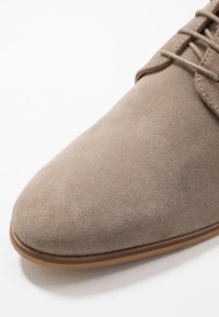Chaussure en daim beige avec un bout pointu, un laçage et une semelle en caoutchouc brun. Texture lisse sans motifs ni accents visibles.