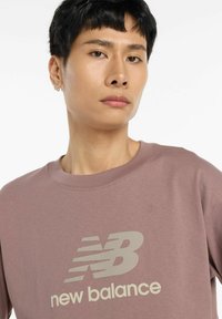 Vaaleanlila puuvillasekoitepaita, jossa on suuri valkoinen "new balance" -logo edessä ja pyöreä kaula-aukko.