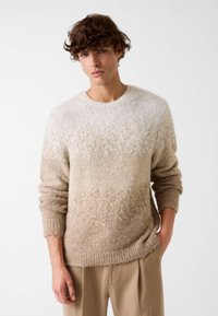 Maglione beige con gradiente e una finitura morbida e testurizzata. Presenta polsini e orlo a costine. Indossato con pantaloni su misura di colore chiaro.