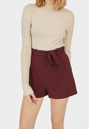 Frau trägt ein beiges, geripptes Langarmshirt, das in hoch taillierte, weinrote Shorts mit einem gebundenen Stoffgürtel gesteckt ist.