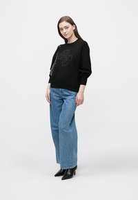 Czarna dzianinowa bluza z wypukłym wzorem, zestawiona z jasnoniebieskimi szerokimi spodniami i czarnymi szpilkami z ostro zakończonym noskiem. Modelka stoi na tle jednolitego tła.