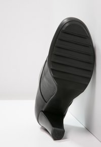 Semelle de chaussure noire à talons hauts avec un fond en caoutchouc texturé, présentant des rainures verticales pour une meilleure adhérence et une légère courbe le long du bord.