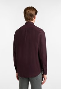 Uomo con capelli castani corti che indossa una maglietta a maniche lunghe di un viola scuro e pantaloni grigi, rivolto di spalle su uno sfondo bianco.