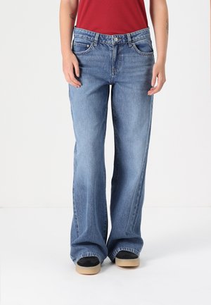 Person iført lyseblå bredbenede jeans og sorte sneakers med beige såler, stående mod en ensfarvet hvid baggrund.