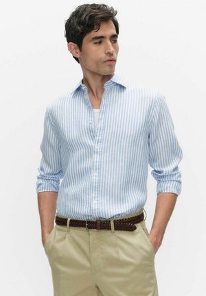 Mand iført lyseblå og hvide stribede skjorte med opklebede ærmer, beige bukser og flettet brunt bælte, hænderne i lommerne.