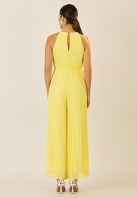 Combinaison jaune plissée avec col haut et dos en trou de clé. Texture lisse, jambes larges, taille cintrée, associée à des talons métalliques.