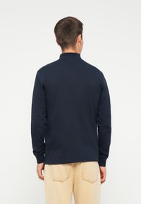 Marineblauwe polo met lange mouwen en hoge kraag, gemaakt van een gladde, gestructureerde stof met geribde manchetten, gedragen met beige broeken.