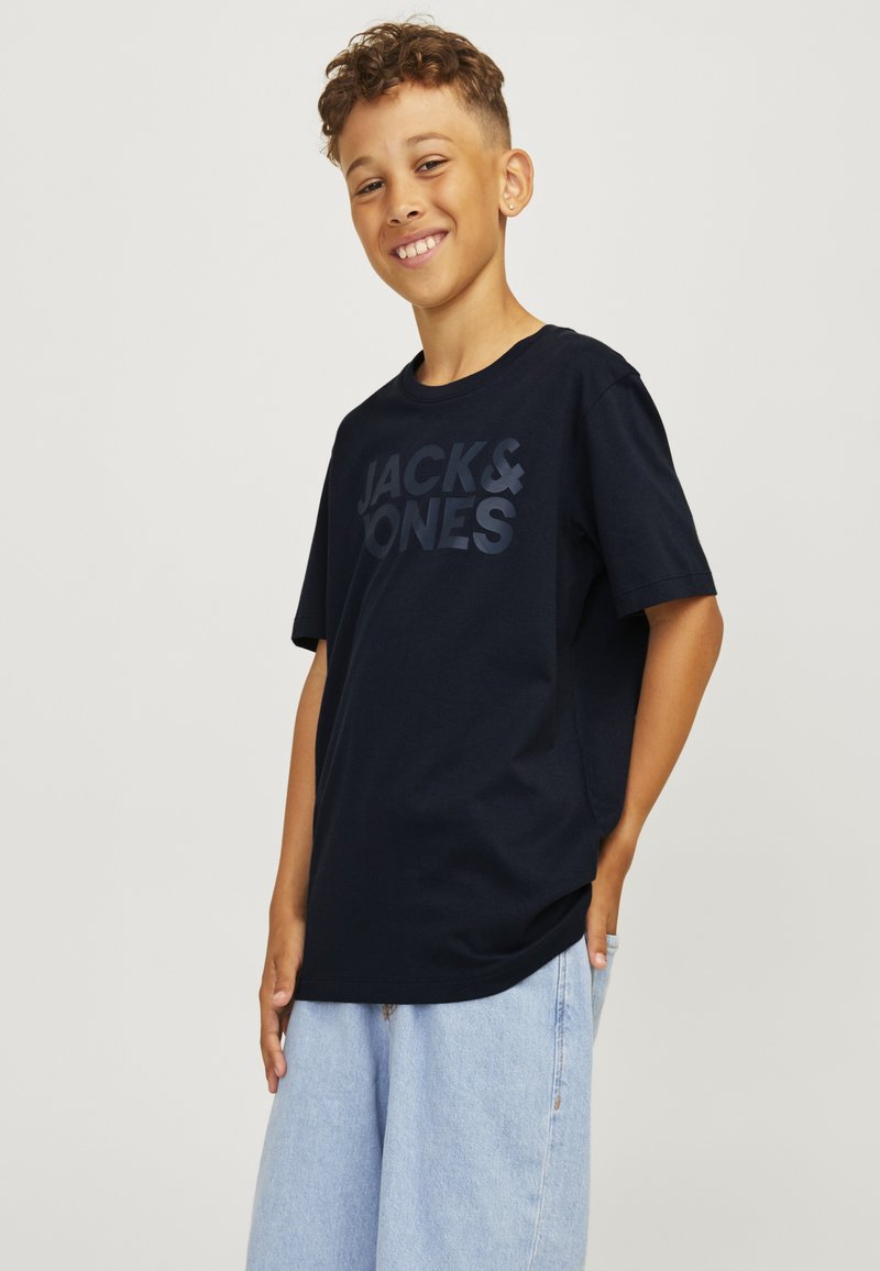 Niño con cabello corto y rizado, vestido con una camiseta negra de Jack & Jones y vaqueros anchos azul claro, sonriendo con una mano en el bolsillo.