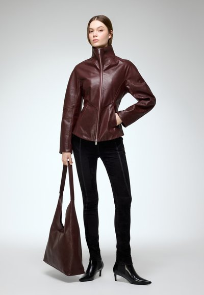 Bordeaux leren jas met hoge kraag en een ritssluiting aan de voorkant. Zwarte fluwelen broek en een grote bijpassende handtas. Model draagt zwarte hoge hakken.