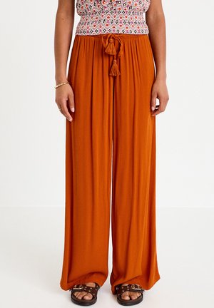 Vrouw draagt roestoranje wijde broek met kwastjes riem, patroonloze mouwloze top en bruine met studs versierde sandalen tegen een witte achtergrond.