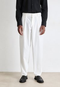 White pleated trousers made of textured fabric, featuring a relaxed fit and paired with black shoes, worn with a black button-up shirt.